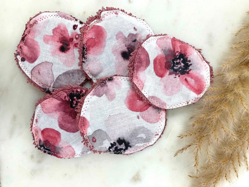 May include: A collection of six round, reusable facial pads. Each pad features a watercolor floral design in shades of pink, gray, and black on a white background. The edges are finished with a ruffled, mauve trim. These pads are designed for skincare.