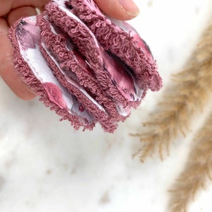 May include: A stack of reusable facial rounds. The rounds are a dusty rose color with a floral pattern on one side and a white fabric on the other. The edges are trimmed with a soft, textured material.