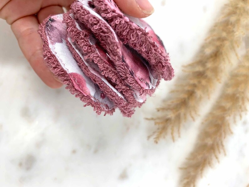 May include: A stack of reusable facial rounds. The rounds are a dusty rose color with a floral pattern on one side and a white fabric on the other. The edges are trimmed with a soft, textured material.