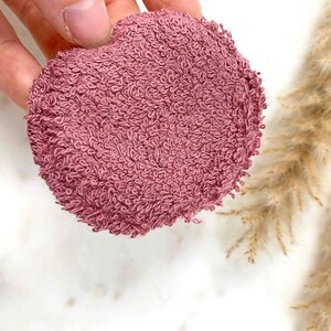 May include: A round, mauve-colored face cleansing pad made of a soft, looped terry cloth material. The pad is held by a hand and is set against a white background with dried ornamental grass.