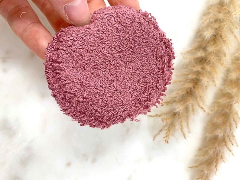 May include: A round, mauve-colored face cleansing pad made of a soft, looped terry cloth material. The pad is held by a hand and is set against a white background with dried ornamental grass.