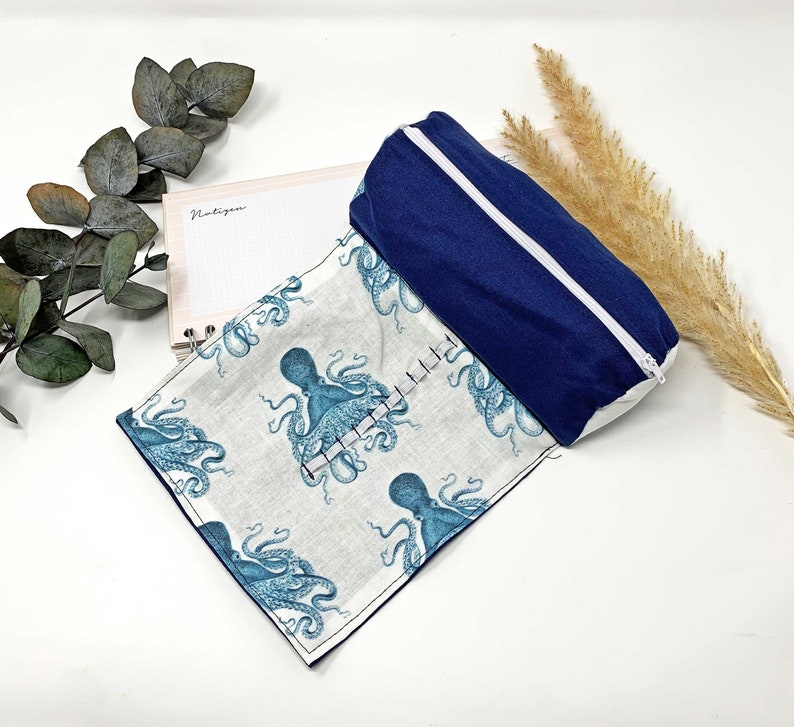 Pode incluir: Um estojo de l&aacute;pis de tecido branco com um z&iacute;per azul e uma estampa de polvo azul. O estojo est&aacute; dobrado ao meio e uma bolsa azul com z&iacute;per est&aacute; em cima do estojo.
