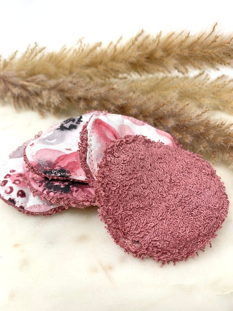 May include: A collection of reusable facial rounds. Some rounds feature a floral pattern in pink, white, and gray, while others are a solid mauve color. The rounds are made of soft, absorbent fabric, ideal for skincare routines.
