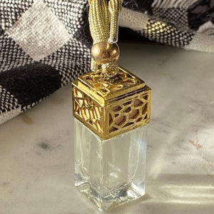 May include: A gold-coloured car air freshener with a geometric design and a tassel. The air freshener is made of glass and has a clear liquid inside.