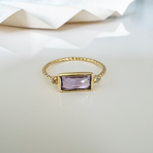 May include: A gold ring with a twisted band and a rectangular purple gemstone. Two smaller clear gemstones are set on either side of the larger gemstone.