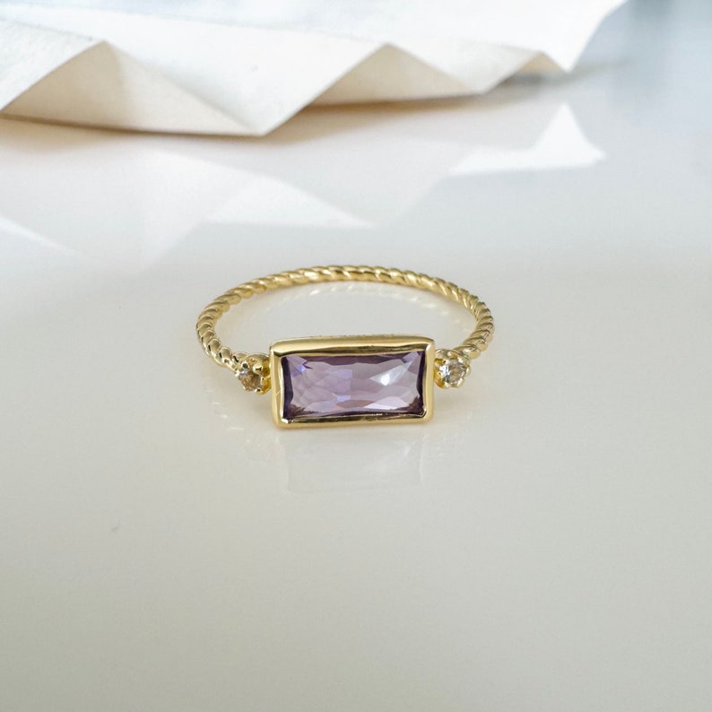 May include: A gold ring with a twisted band and a rectangular purple gemstone. Two smaller clear gemstones are set on either side of the larger gemstone.