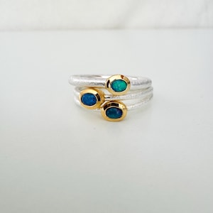 Può includere: Tre anelli d'argento con castoni dorati, ciascuno con una pietra preziosa ovale. Le pietre preziose sono in tonalità blu e verde. Gli anelli sono impilati, creando un effetto a strati. Su sfondo bianco.