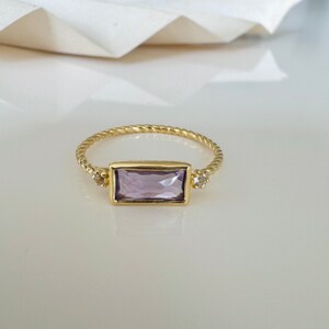 May include: A gold ring with a twisted band and a rectangular purple gemstone. Two smaller clear gemstones are set on either side of the larger stone.