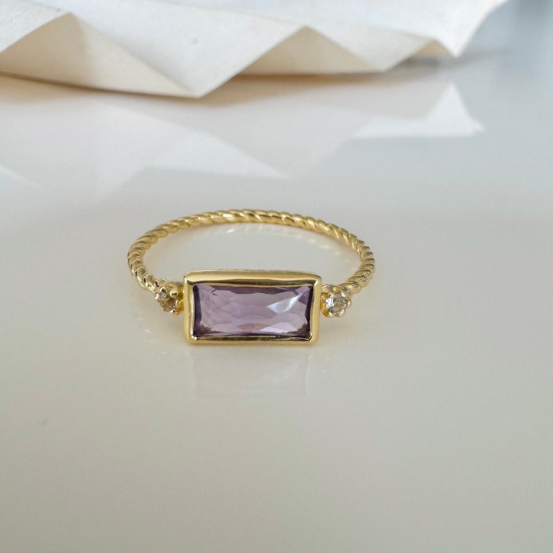 May include: A gold ring with a twisted band and a rectangular purple gemstone. Two smaller clear gemstones are set on either side of the larger stone.