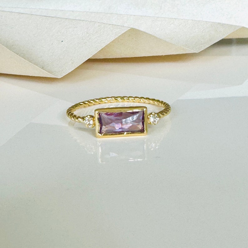 May include: A gold ring with a twisted band and a rectangular purple gemstone set in the center. Two small round white gemstones are set on either side of the center stone.