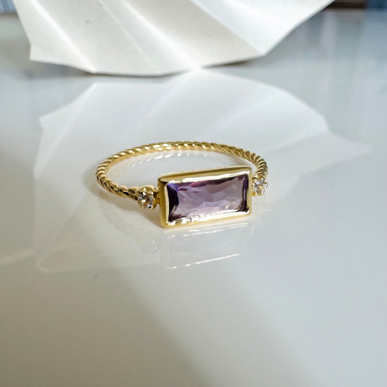 May include: A gold ring with a rectangular amethyst gemstone and two small diamond accents. The ring has a twisted band.