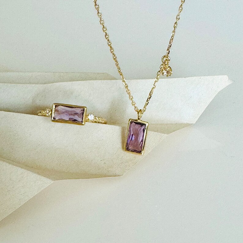 May include: A delicate gold necklace and ring set with a rectangular amethyst gemstone. The necklace features a simple chain and a pendant with a single amethyst stone. The ring has a thin band with a single amethyst stone.