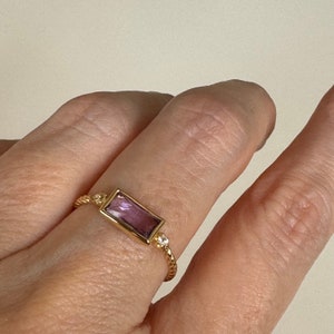 May include: A gold ring with a rectangular purple gemstone and a small clear gemstone. The ring has a twisted band.