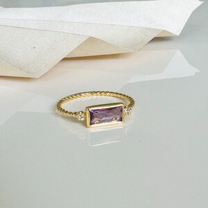 May include: A gold ring with a twisted band and a rectangular purple gemstone set in the center. Two smaller round gemstones are set on either side of the larger stone.