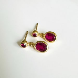 Könnte beinhalten: Ein Paar goldfarbene Ohrhänger mit ovalen, tiefroten Edelsteinen. Jeder Ohrring hat einen kleinen, runden Edelstein oben, der durch eine goldene Kette mit einem größeren, ovalen Edelstein verbunden ist. Auf weißem Hintergrund.