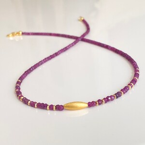 Könnte beinhalten: Eine zarte goldene Halskette mit einem kleinen goldenen Barren-Anhänger und winzigen violetten Edelsteinperlen.