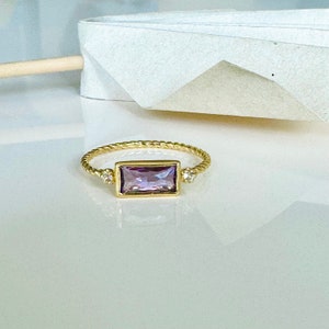 May include: A gold ring with a twisted band and a rectangular purple gemstone. Two smaller clear gemstones are set on either side of the larger stone.