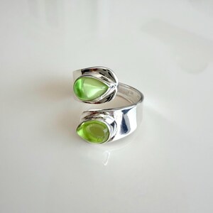 Könnte beinhalten: Ein offener Ring aus poliertem Silber mit zwei tropfenförmigen, hellgrünen Cabochon-Steinen. Das Design des Rings lässt die beiden Steine einander gegenüberliegen, was für ein symmetrisches Aussehen mit einer glatten, reflektierenden Oberfläche sorgt.
