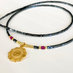 Pode incluir: Um colar de ouro com um pingente de design circular detalhado pendurado em uma corrente de contas pretas e douradas. A corrente é em camadas e apresenta pequenas contas vermelhas.