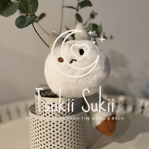 May include: White plush toy shaped like a moon with a heart and a face. The toy is sitting in a white metal container with holes. The text "Tsukii Suki" is written on the container. The text "Gifts with love from the moon & back" is written below the container.