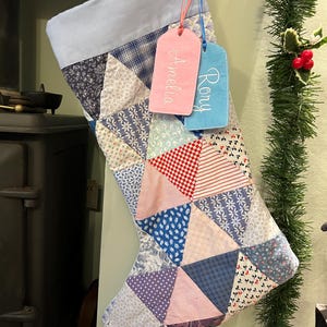May include: A patchwork Christmas stocking in a variety of fabrics and colours, including blue, red, and white. The stocking has a light blue cuff and is decorated with two tags, one pink and one blue, embroidered with the names "Amelia" and "Rory".