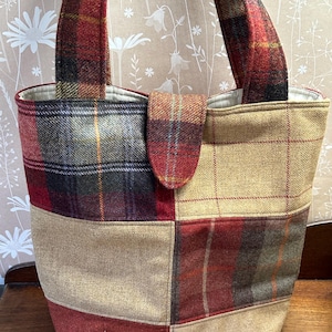 May include: A patchwork tote bag in a tartan pattern, featuring red, beige, and dark green hues. The bag has two handles and a flap closure. The material is a thick, textured fabric.
