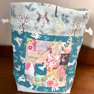 May include: A drawstring bag with a patchwork design. The bag has a teal border with white rabbits and a light blue top with a forest scene. The patchwork section features floral and rabbit patterns.