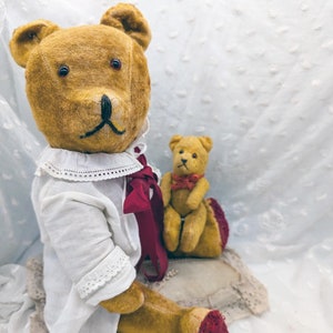 Könnte beinhalten: Zwei Vintage-Teddybären, einer trägt ein weißes Kleid mit einem roten Band, sitzen auf einem weißen Spitzenstoff. Der größere Bär hat braune Augen und eine braune Nase. Der kleinere Bär hat braune Augen und eine braune Nase.