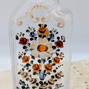 Könnte beinhalten: Transparente Glasflasche mit silbernem Metallverschluss, verziert mit handgemalten Blumenmustern. Die Blumen sind in Rot-, Orange-, Blau- und Weißtönen gehalten und erzeugen eine Vintage-Ästhetik. Die Flasche ist rechteckig mit abgerundeten Kanten.