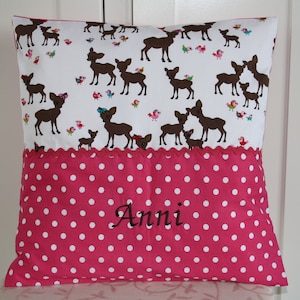 Puede incluir: Una almohada rosa y blanca con un patrón de lunares blancos. La parte delantera de la almohada tiene una tela blanca con un patrón de ciervos marrones y el texto "Anni" bordado en negro.