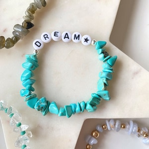 Puede incluir: Pulsera de piedras de chip de turquesa con cuentas de letras blancas que deletrean "DREAM" y un dije de estrella.