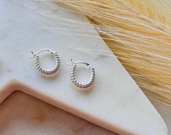 May include: A pair of small, silver hoop earrings. Each earring has a textured, dotted design around the hoop. The earrings are displayed on a white marble surface with a neutral background.