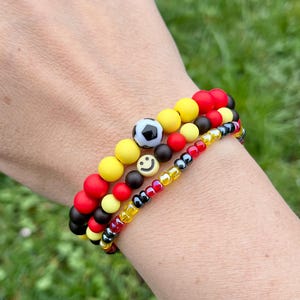Könnte beinhalten: Drei Armbänder mit roten, gelben, schwarzen und weißen Perlen. Ein Armband hat eine Fußballperle, ein anderes eine Smiley-Gesicht-Perle. Die Armbänder sind an einem Handgelenk gestapelt.