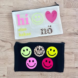Könnte beinhalten: Eine weiße Reißverschlusstasche mit einem neonpinken 'hi', einem neongelben Smiley, einem pinken Herz sowie dem Text 'ciao kakao' und 'nö' in Leopardenmuster. Darunter eine schwarze Reißverschlusstasche mit fünf Smileys in Neongelb, Leopardenmuster und Pink.