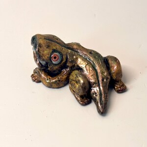 May include: A handcrafted frog figurine with a detailed, textured surface. The frog is primarily bronze and green with red and white eyes. The figurine is posed in a resting position, showcasing its legs and body.