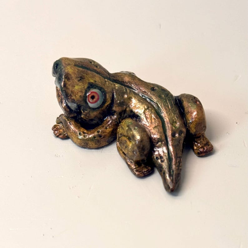 May include: A handcrafted frog figurine with a detailed, textured surface. The frog is primarily bronze and green with red and white eyes. The figurine is posed in a resting position, showcasing its legs and body.