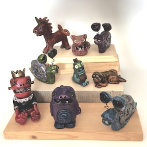 May include: A collection of whimsical, handmade ceramic figurines. The figures include a unicorn, various monsters, and a snail, each with unique features and vibrant colors. They are displayed on a wooden tiered stand, showcasing their detailed craftsmanship.