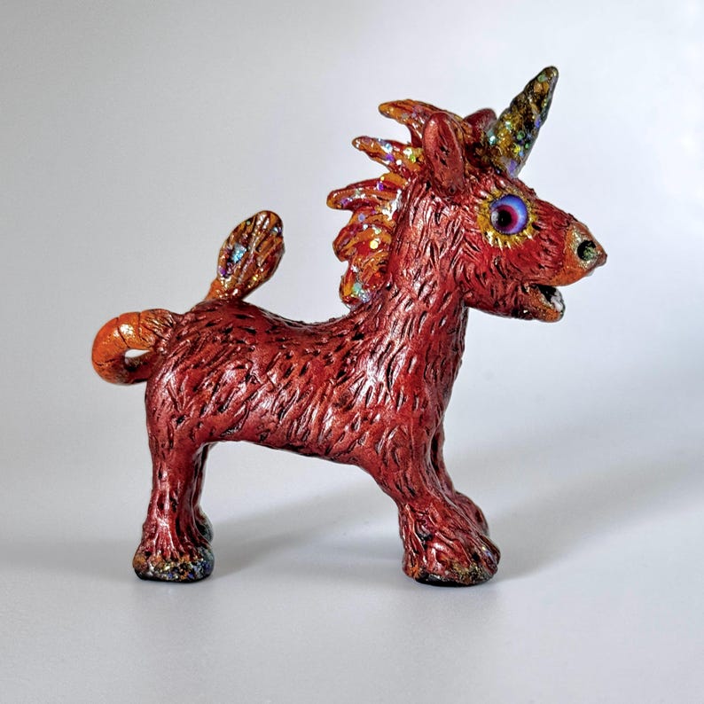 May include: A handcrafted, red-toned unicorn figurine with a textured surface. The mythical creature features a glittery mane, tail, and horn, with iridescent accents on its wings and eyes. The unicorn is posed on all fours, showcasing its unique design.