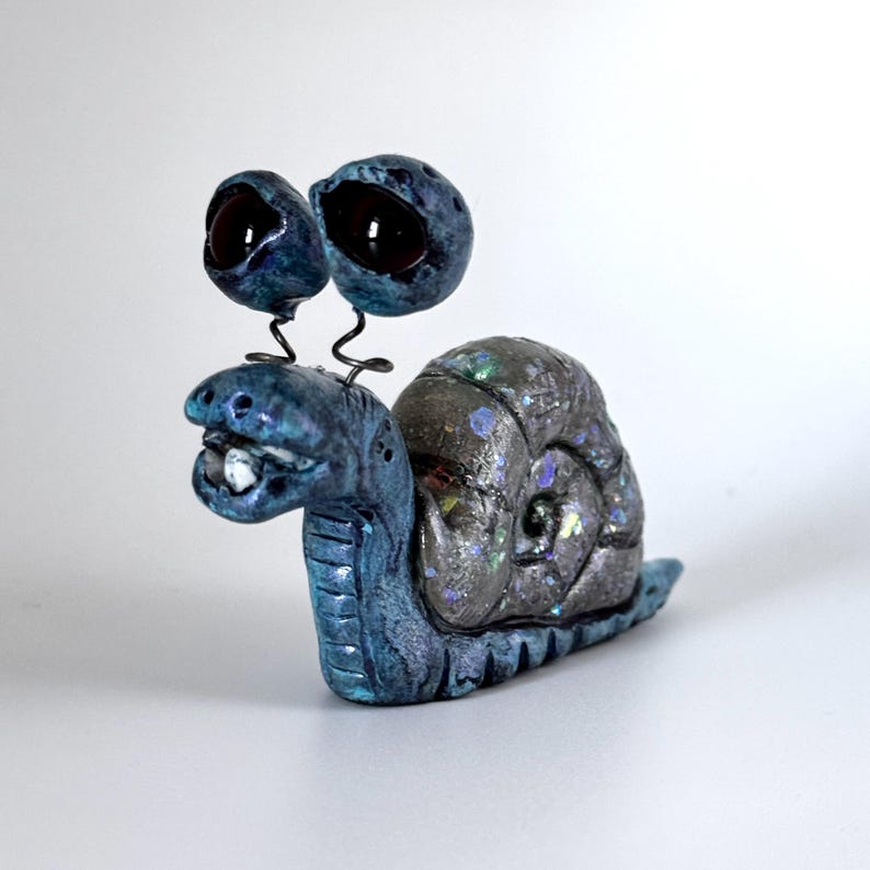 May include: A whimsical, handmade snail figurine with large, bulbous eyes and a textured shell. The snail's body is a gradient of blue hues, and the shell is a mix of gray and iridescent colors. The eyes are dark, and the snail has a smiling expression.