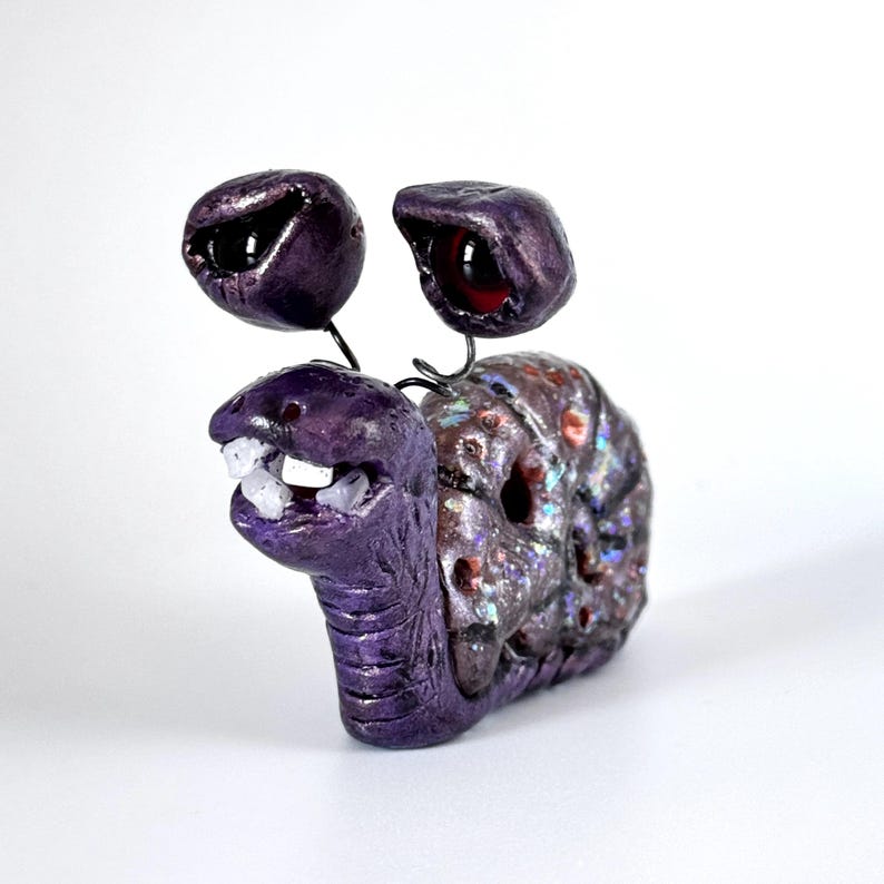 May include: A whimsical, handmade sculpture of a snail with large, bulbous eyes and a toothy grin. The snail's body is a deep purple, with a textured shell in shades of purple, brown, and iridescent accents. The eyes are black and red.