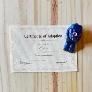 May include: A blue figurine of a creature with large eyes rests next to a certificate of adoption. The certificate is for "Sylvia" and is dated 12/30/2025. The figurine has a glossy finish and is on a light wood surface.