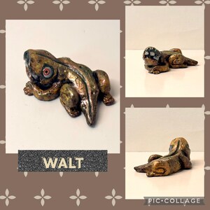 May include: A handcrafted, bronze-colored frog figurine with red eyes. The frog is in a resting pose, with detailed texture and a metallic finish. The name "WALT" is displayed on a rectangular label below the frog.
