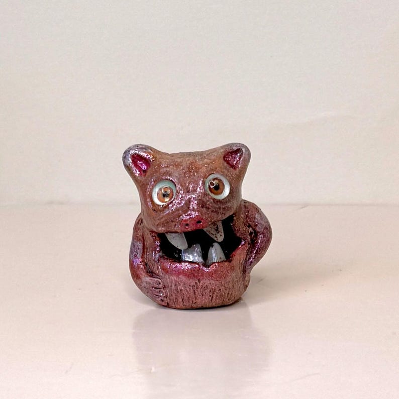 May include: A small, brown, monster figurine with a wide, toothy grin and large, round eyes. The figurine has a textured surface and a metallic sheen, with purple accents around the ears and mouth. The creature appears to be a handmade art piece.