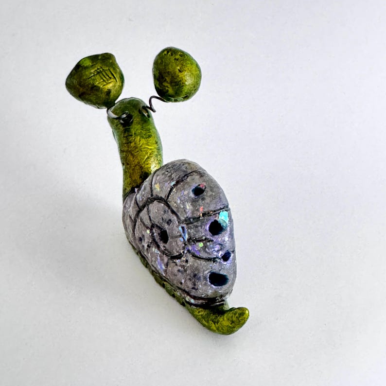 May include: A whimsical, handmade snail figurine. The snail features a green body and head, a gray shell with iridescent accents, and two green, rounded antennae. The figurine is set against a white background.