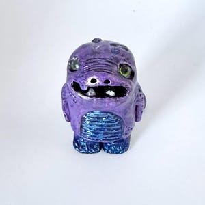 May include: A whimsical, purple creature figurine with a textured surface and a friendly expression. The figurine has green eyes, a silver-colored mouth, and blue accents on its belly and feet. It is a unique, handmade art piece.