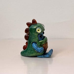 May include: A small, handcrafted figurine of a whimsical creature. The figure is primarily green with brown, blue, and yellow accents. It has large, round eyes and a friendly expression, suggesting a fantasy or collectible item.