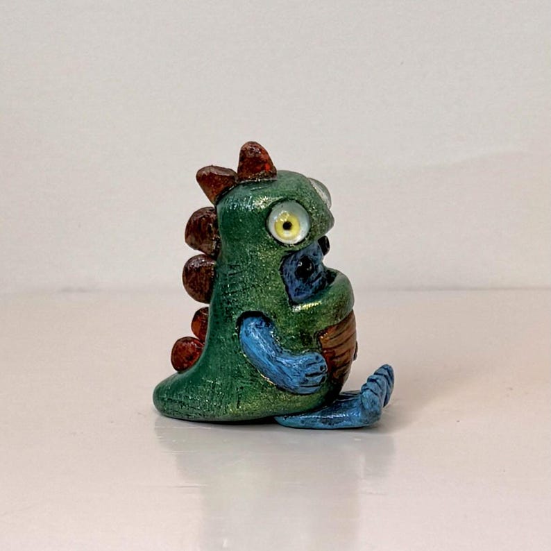 May include: A small, handcrafted figurine of a whimsical creature. The figure is primarily green with brown, blue, and yellow accents. It has large, round eyes and a friendly expression, suggesting a fantasy or collectible item.