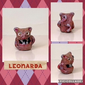 May include: A handmade, reddish-brown ceramic figurine with a wide open mouth revealing teeth. The figurine has large, round eyes and is labeled "LEONARDA" in a banner. The background is a collage with a diamond pattern.