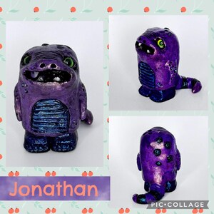 May include: A handcrafted purple monster figurine with green eyes, a toothy grin, and a blue belly. The figurine is painted with metallic accents and has a small tail. The name "Jonathan" is written on a purple banner below the figurine.