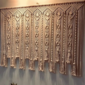 Puede incluir: Un gran tapiz de macramé beige. La pieza de arte textil presenta intrincados patrones de nudos, incluyendo formas de diamante y hebras verticales con borlas. Está montado sobre una barra de madera y cuelga de una pared gris claro.